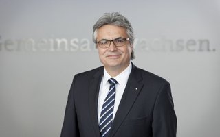 Portrait eines Mannes mittleren Alters mit grauen Haaren und heller Hautfarbe. Er trägt eine Brille und einen Anzug.