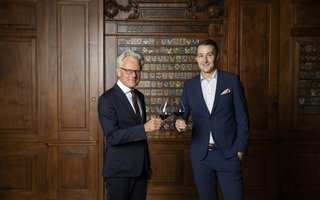 Zwei Männer in Anzügen stehen vor einer dunkelbraunen Holzwand und stossen mit einem Glas Rotwein an.