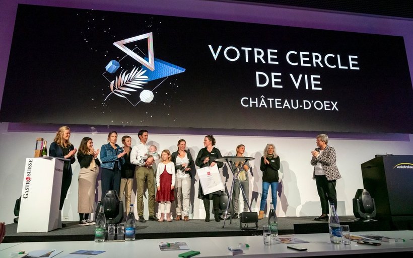 13 Personen stehen auf einer Bühne, dahinter befindet sich eine grosse Leinwand. Auf ihr wird der Name der Gewinner vom Hotel-Innovations-Tag abgebildet.